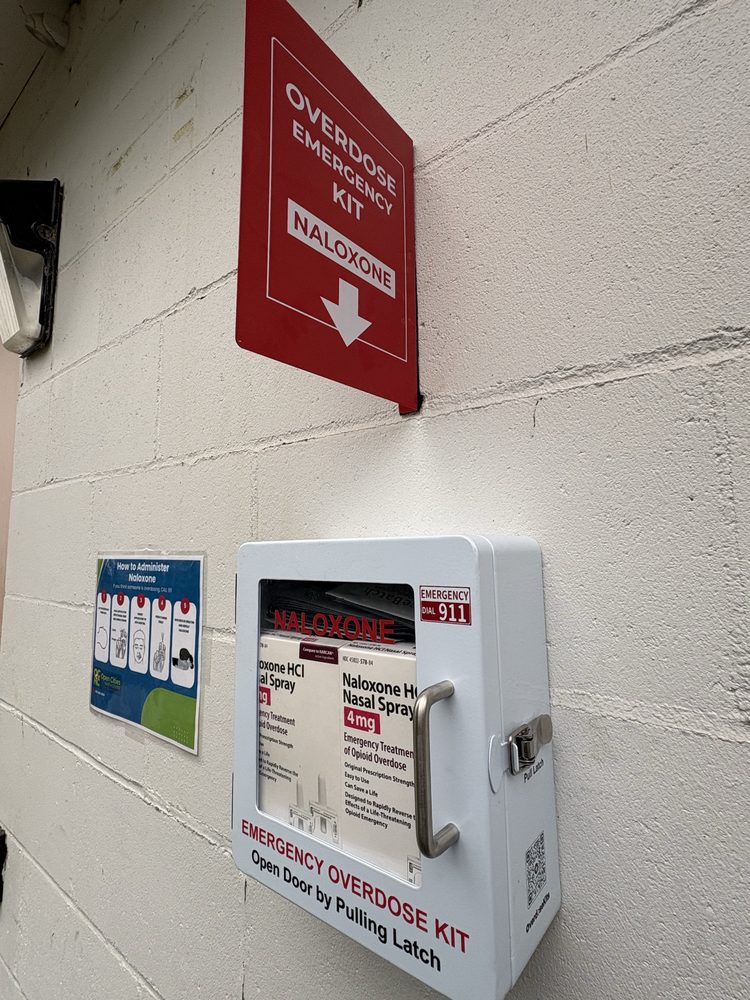 Outdoor Emergency Overdose Kit box stocked with naloxone nasal spray, mounted on the wall behind the Open Cities Health Center North End Clinic